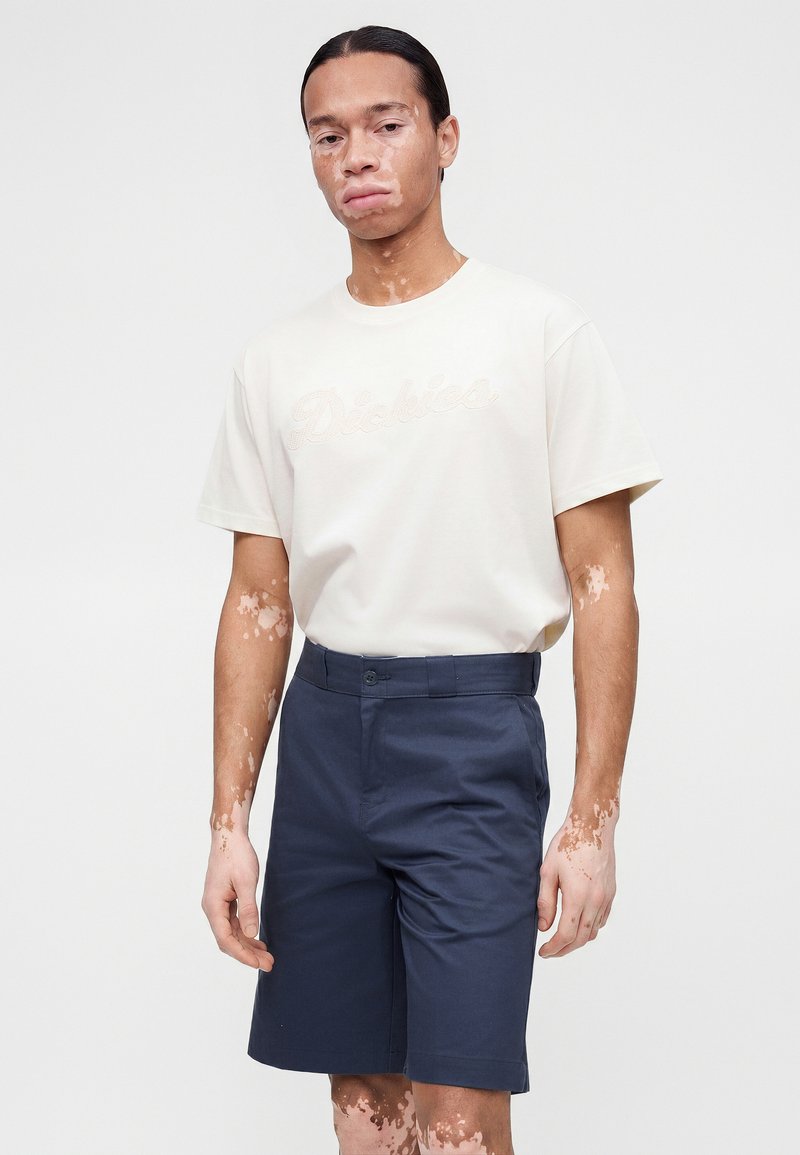 Mann mit Vitiligo, der ein weißes kurzärmeliges Hemd trägt, das in marineblaue knielange Shorts gesteckt ist, steht vor einem einfarbigen Hintergrund.