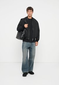 Sort pufferjakke over en sort skjorte, parret med løse, lyseblå jeans og sorte sko. Modellen holder en sort taske med strukturerede detaljer.