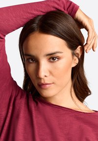 Bordeauxrotes Langarmshirt mit einem lässigen Halsausschnitt. Das Model hat glattes, dunkelbraunes Haar und minimales Make-up, mit heller Haut und Ohrringen.