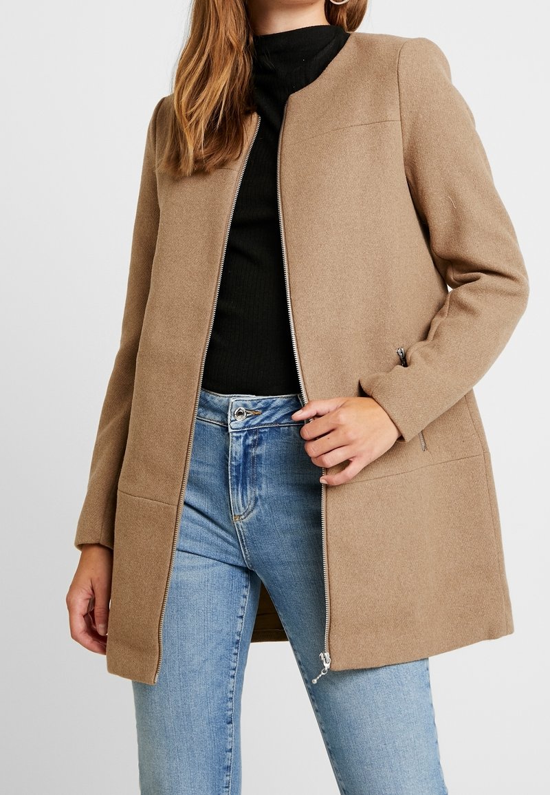 Hnědý saka střižený na míru s předním zipem, dlouhými rukávy, bočními kapsami a hladkým povrchem. Doplňuje ho modré džíny.