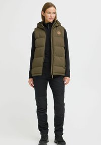 Gilet rembourré vert olive avec capuche, devant zippé et patch logo, associé à un pantalon noir et des bottes, présentant une finition lisse et texturée.