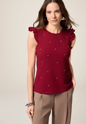 Haut sans manches bordeaux avec des accents de volants aux épaules, orné de petits clous en argent, associé à un pantalon beige.
