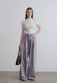 Φαρδιά παντελόνια ανοιχτού γκρι χρώματος με ψηλή μέση, συνδυασμένα με εφαρμοστό λευκό ριμπ μπλουζάκι. Το μοντέλο κρατάει μια σκούρα τσάντα.