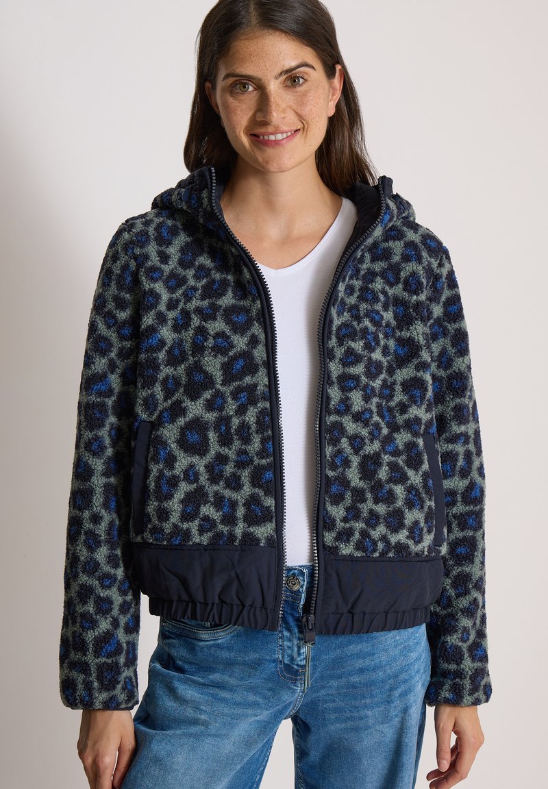 Jeune femme aux longs cheveux noirs porte une veste polaire bleue et noire à imprimé léopard sur une chemise blanche et un jean bleu, souriant légèrement.