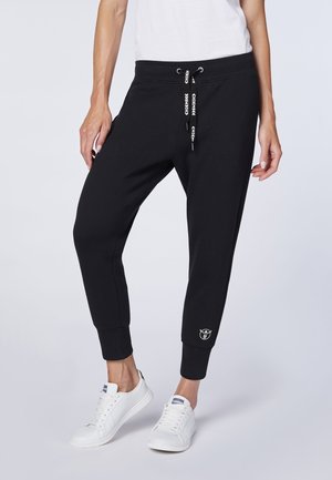 Personne portant un pantalon de jogging noir avec des cordons de serrage de marque et des baskets blanches, debout devant un fond clair uni.