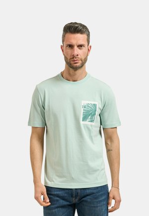 Hombre con cabello corto y barba, lleva una camiseta verde claro con gráfico de hoja de palma en el bolsillo del pecho y vaqueros azules, de pie frente a un fondo liso.