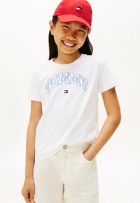 Hvid t-shirt med blå "TOMMY" bogstaver, parret med lysebeige bukser, suppleret af en rød kasket med et lille logo.