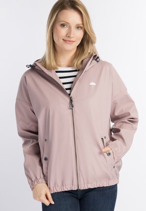 Giovane donna che indossa una giacca da pioggia rosa chiaro con zip e cappuccio e una maglietta a righe sotto, in piedi con una mano in tasca.