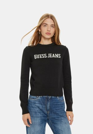 Maglione nero a maglia con orlo e polsini a coste, caratterizzato da testo bianco "GUESS JEANS" sulla parte anteriore. Abbinato a jeans blu.