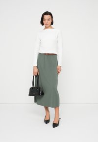 Λευκό μακρυμάνικο τοπ, πράσινη μάξι φούστα, καφέ ζώνη, μαύρη τσάντα χειρός και μυτερά μαύρα ψηλοτάκουνα. Λείο ύφασμα, εφαρμοστή σχεδίαση και ουδέτερα χρώματα.