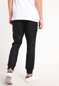 Person med svarta joggingsbyxor och vita Nike-sneakers, som står med ryggen mot kameran framför en enfärgad vit bakgrund.