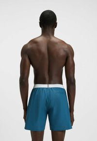 Uomo muscoloso visto di spalle che indossa pantaloni da bagno blu con vita elastica bianca su uno sfondo chiaro e semplice.