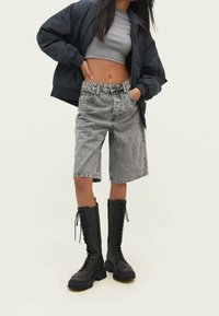 Grijze denim Bermuda-shorts met een losse pasvorm, hoge taille en contrasterende stiksels, gecombineerd met kniehoge zwarte veterschoenen en een donkere jacket.