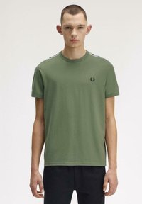 Jeune homme aux cheveux courts portant un t-shirt vert olive avec un logo noir sur la poitrine et un pantalon noir, sur un fond clair uni.