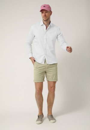 Hombre con gorra rosa, camisa blanca abotonada, pantalones cortos beige y zapatos grises sin cordones, de pie con una mano en el bolsillo.