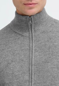 Szary sweter na zamek błyskawiczny, wykonany z teksturowanej tkaniny, z wysokim kołnierzem. Posiada frontowy zamek z dopasowaną zawieszką oraz prążkowane akcenty.