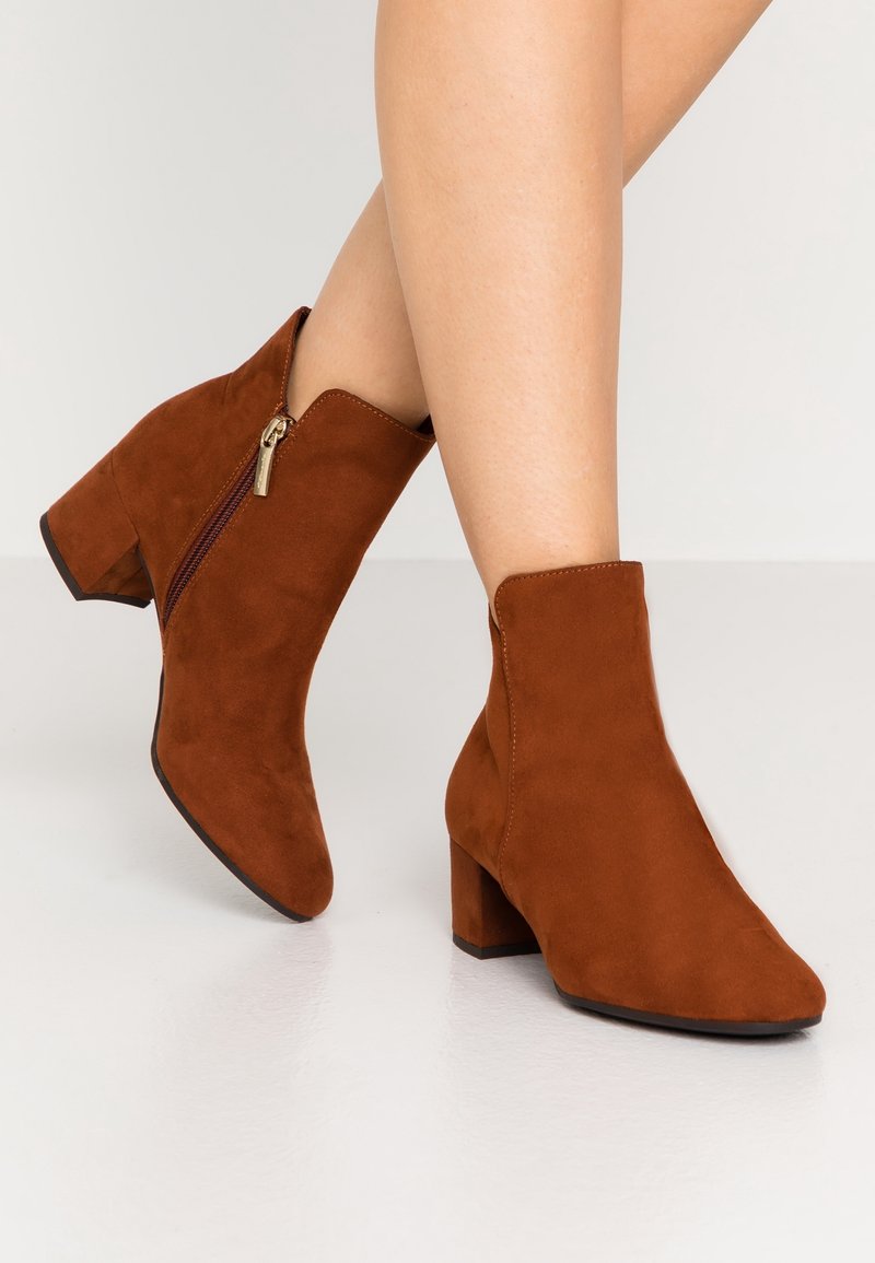 Bottines en daim marron à talons blocs et fermetures éclair latérales portées sur des jambes croisées sur un fond uni clair.