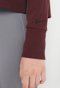 Chemise à manches longues bordeaux à texture côtelée, avec un logo Nike noir sur le poignet et un vêtement gris ajusté en dessous.
