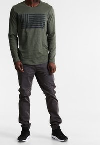 Olijfgroene shirt met lange mouwen en een marineblauwe horizontale gestreepte vlaggrafiek. Gecombineerd met grijze taps toelopende broek en zwarte sneakers.