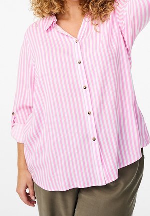 Žena v růžové a bílé svisle pruhované košili s knoflíky a vyhrnutými rukávy, a v olivově zelených kalhotách, stojící před bílým pozadím.