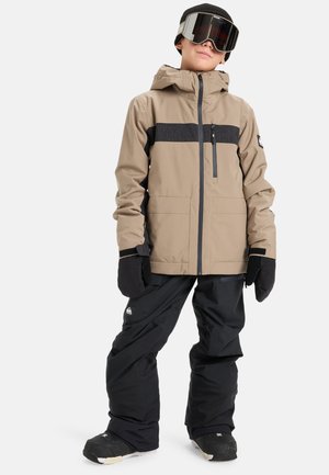 Enfant portant une veste de neige tan et noire, un pantalon de neige noir, des gants noirs, un bonnet noir et des lunettes de ski se tenant contre un fond blanc.