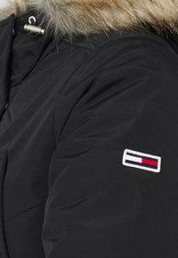 Zwarte gewatteerde jas met een kraag van bont, met een geborduurd logo in rood, wit en blauw op de mouw.