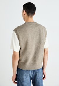 Un pull sans manches en tricot beige avec un col rond, associé à une chemise blanche à manches courtes, porté avec un jean en denim bleu.