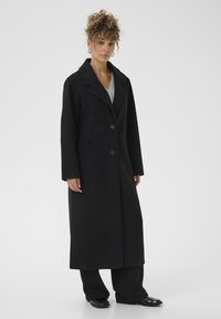 Woman with curly hair wears long black coat over gray top and black pants, standing against a plain white background.