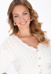 Fehér kötött kardigán struktúrált mintával, V-nyakkivágással és díszítő gombokkal. Csillogó fülbevalókkal párosítva.