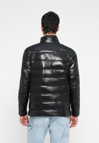 Juoda pūkinė striukė su aukštu apykakliu, su siuvinėtu dizainu, blizgia apdaila ir logotipu gale. Dėvima su šviesiai mėlynomis džinsais.