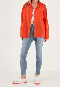 Femme portant une chemise surdimensionnée orange fluo, un crop top blanc, un jean skinny bleu clair et des baskets blanches avec des semelles orange.