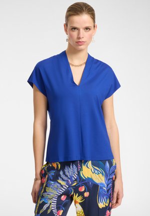 Top blu fitted a maniche corte con colletto a V. Texture liscia, design casual. Abbinato a pantaloni con motivo a fantasia che presentano colori vivaci e motivi.