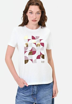 Donna con una maglietta bianca con motivo geometrico in bordeaux, oro e semicerchi punteggiati, abbinata a jeans di jeans blu.