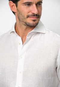 Camisa de lino blanca con cuello abotonado, textura sutil y cuatro botones en la parte delantera. Tejido suave con un ajuste ligeramente relajado.