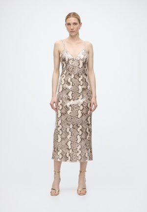 Femme portant une robe sans manches, col en V, longueur cheville imprimé serpent, fines bretelles et talons à bride dorée à la cheville, debout devant un fond blanc uni.