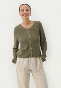Olivgrüner gerippter Cardigan mit Rundhalsausschnitt, langen Ärmeln und Knopfleiste, kombiniert mit hellen, locker sitzenden Hosen.