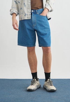 Άτομο που φοράει μπλε τζιν σόρτς, καφέ πουκάμισο, ανοιχτόχρωμο τζάκετ tie-dye, μαύρες κάλτσες και ανοιχτό γκρι αθλητικά παπούτσια, στέκεται σε μπλε πάτωμα.