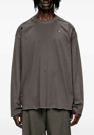 Persona che indossa una maglia grigia a maniche lunghe, larga, con cuciture visibili e un piccolo logo rosso vicino alla spalla sinistra, abbinata a pantaloni grigi.