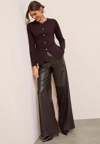 Bordo kardigāns ar zeltītām pogām, asimetriska apakšmala, apvienots ar platām brūnām zamša biksēm un virsotnēm ar spiciem burgundijas krāsas papēžiem. Redzamas gludās tekstūras.