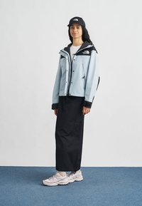 Αδιάβροχο μπουφάν γαλάζιου χρώματος με μαύρες λεπτομέρειες, μαύρο παντελόνι και μαύρο καπέλο. Διαθέτει φερμουάρ εμπρός και τσέπες· φορεμένο με αθλητικά παπούτσια ανοιχτού χρώματος.