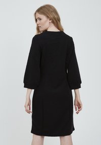 Musta mekko, jossa on kolmivuotiset hihat, teksturoitu kangas, istuva siluetti ja pyöreä pääntie; yksinkertainen ja tyylikäs malli, jossa ei ole näkyviä koristuksia.