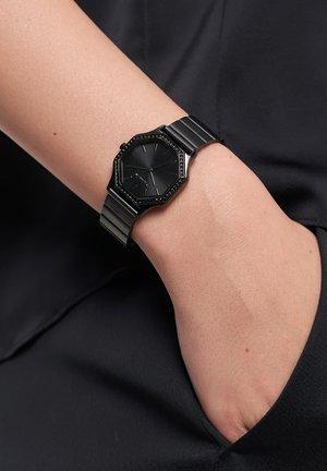 Schwarze achteckige Armbanduhr mit glattem Gehäuse, strukturiertem Armband und minimalistischem schwarzen Ziffernblatt; auf einem Handgelenk vor einem dunklen Hintergrund präsentiert.