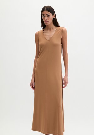 Frau steht und trägt ein ärmelloses, kamelfarbenes, bodenlanges Kleid mit V-Ausschnitt vor einem schlichten weißen Hintergrund.