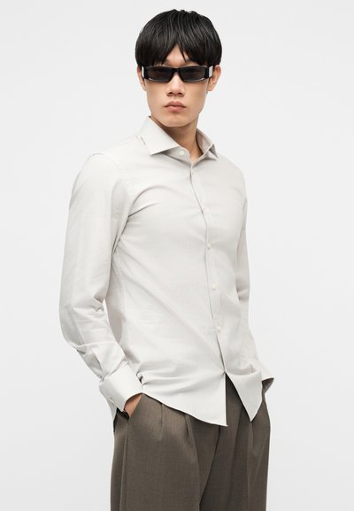 Νεαρός άνδρας που φορά ανοιχτό γκρι πουκάμισο με κουμπιά, καφέ πιετέ παντελόνι και σκούρα ορθογώνια γυαλιά ηλίου, με τα χέρια στις τσέπες.