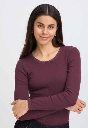 Ung kvinde med langt mørkt hår iført en tætsiddende lilla langærmet top med blondekant, smilende med armene krydset mod en ensfarvet baggrund.