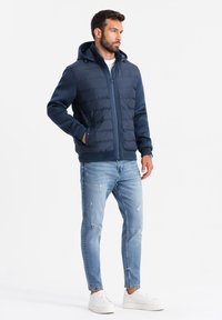 Marinblå puffjacka med huva, med en kviltad övre del, kontrasterande stickade ärmar och dragkedja. Kombinerad med ljusblå slitna jeans.