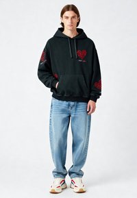 Melns kapuci ar sarkaniem sirds rakstiem, kanguru kabata, saistāmā kapuce. Kombinēts ar gaiši zilu plašu džinsu un daudzkrāsainām sporta apaviem.