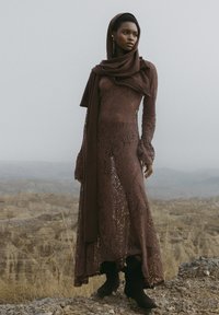 Dlhé hnedé čipkované šaty s ladiacou šatkou, so širokými rukávmi a rozšírenými manžetami, zasadené do drsnej krajiny.