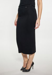 Schwarzer gerippter Strick-Midirock mit einem taillierten Bund, glatter Textur und minimalistischen Design, kombiniert mit spitzen schwarzen High Heels.