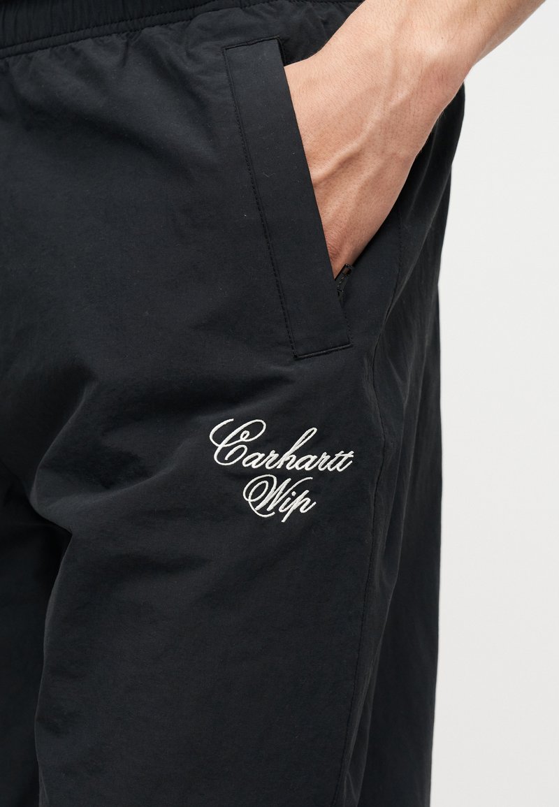 Nahaufnahme einer Hand in der Seitenasche einer schwarzen Carhartt WIP-Hose mit weiß gesticktem Logo am Oberschenkel.
